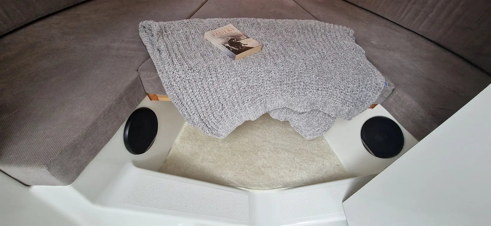 A soft grey blanket and a book lie on a boat's seating area, with round speakers and a white panel visible.